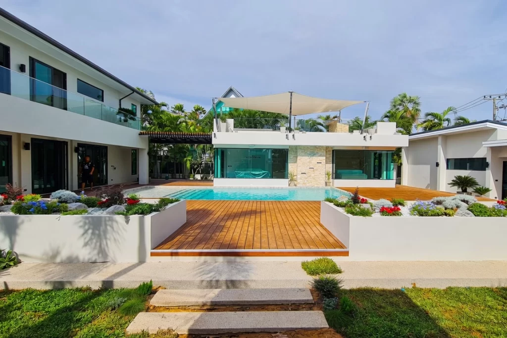 Beautifully renovated modern pool villa in Jomtien, Pattaya featuring a sleek white contemporary architecture, private swimming pool, expansive natural wooden deck, and lush tropical landscaping with white planters filled with vibrant flowers.