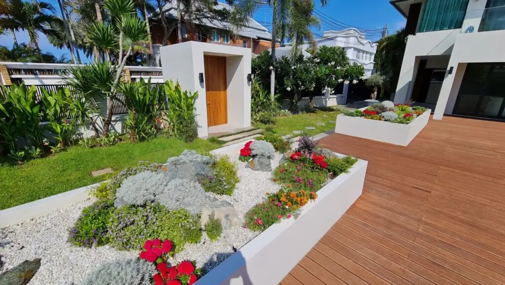 Stunning modern tropical landscape of a luxury villa in Jomtien, Pattaya featuring natural bamboo-style wooden decking, symmetrical white planters with vibrant landscaping, lush palm trees, and a minimalist white entrance pavilion with warm wooden door.