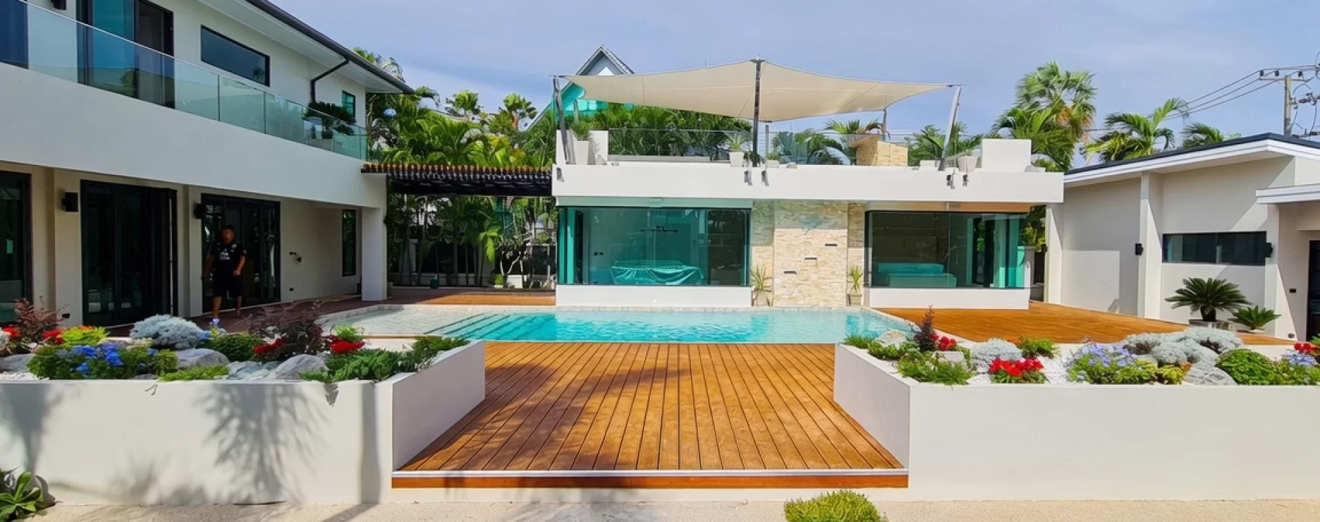 Beautifully renovated modern pool villa in Jomtien, Pattaya featuring a sleek white contemporary architecture, private swimming pool, expansive natural wooden deck, and lush tropical landscaping with white planters filled with vibrant flowers.